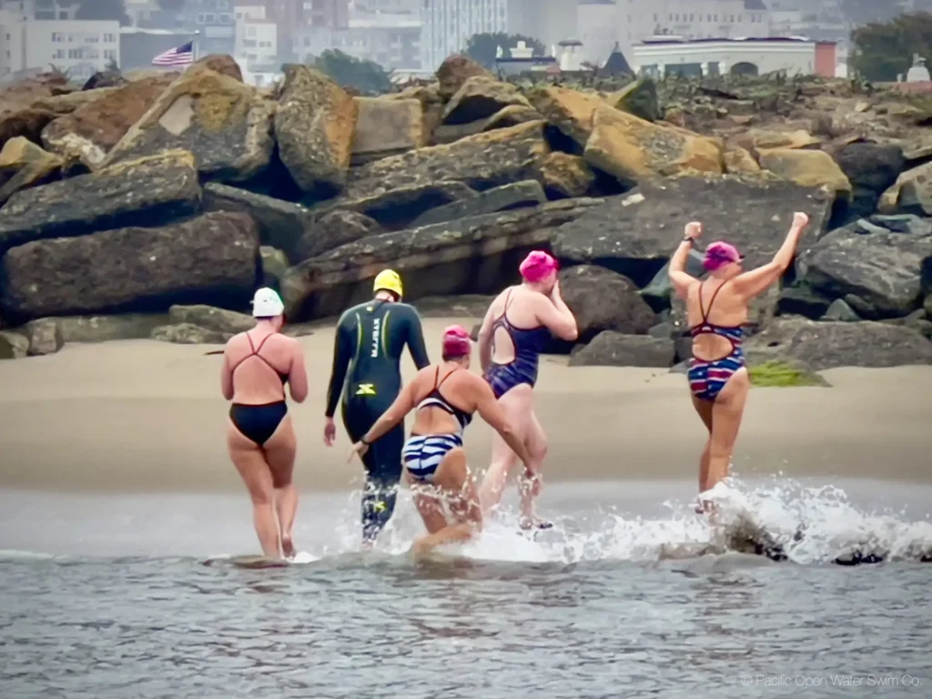 Five swimmers, including one in a wetsuit and others in swimsuits and pink caps, walk triumphantly out of the water toward shore after completing an Alcatraz to San Francisco swim.