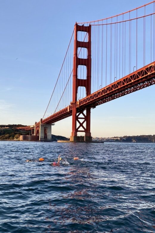 Swimmers in open water under the Golden Gate Bridge, making their way through the San Francisco Bay with bright swim caps and buoys for visibility.